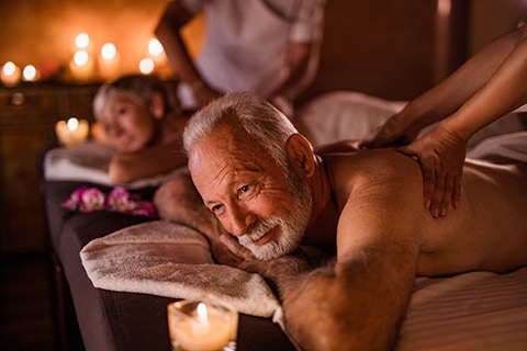 An elderly man and woman in the background getting back massages in a spa room with candles around.