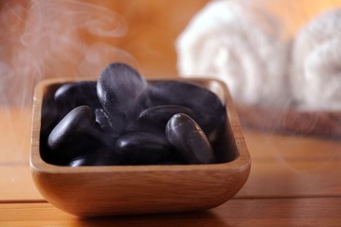 A rustic wooden bowl filled with steaming hot black massage stones sits on a wooden table next to two white towels.